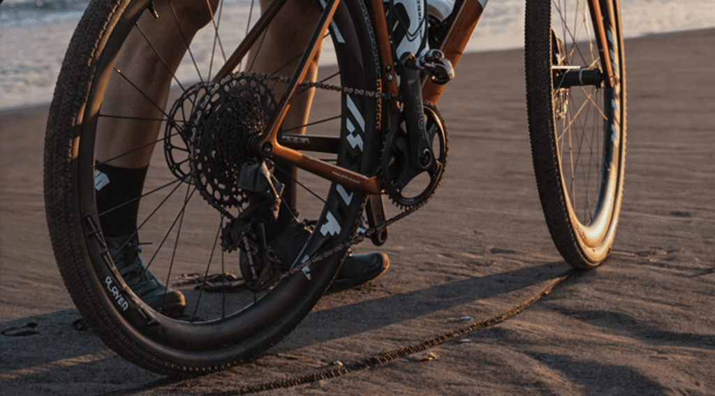 Bicicleta equipada con cambios de bicicleta de alta precisión, detenida en la arena con el mar de fondo. La transmisión luce impecable, con un cassette amplio y un desviador trasero listo para afrontar cualquier terreno. La imagen, tomada al atardecer, resalta la estética y funcionalidad de los componentes. El ciclista, con calcetas y equipamiento técnico, representa la pasión por el ciclismo y la aventura. 